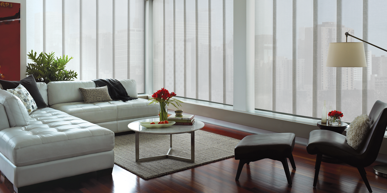 Living room with a sectional sofa, coffee table, and lounge chair surrounded by large windows with panel shades.